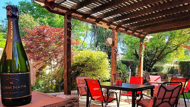 Une terrasse avec des sièges extérieurs, une bouteille de vin Mumm Napa Brut Prestige et une structure en forme de pergola.