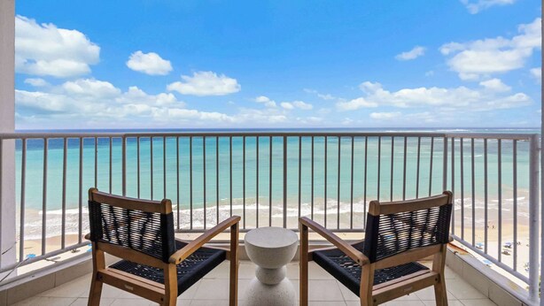 Un balcone con due poltrone in vimini e un tavolino, con vista su una spiaggia di sabbia e acque turchesi cristalline.