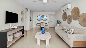 Un soggiorno moderno con TV a schermo piatto, ventilatore a soffitto, zona pranzo con tavolo e sedie, e divano con cuscini decorativi.
