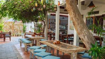 An outdoor seating area with wooden tables and benches, surrounded by potted plants and a tree.