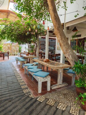 Un espace extérieur aménagé avec des tables et des bancs en bois, entouré de plantes en pot et d’un arbre.
