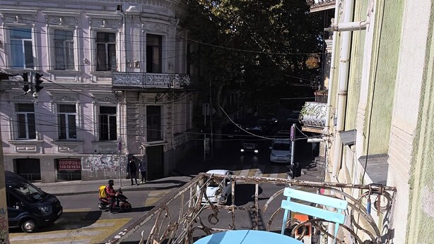 Un balcón con una mesa y sillas azules, vista de una calle con un edificio al fondo y un árbol.