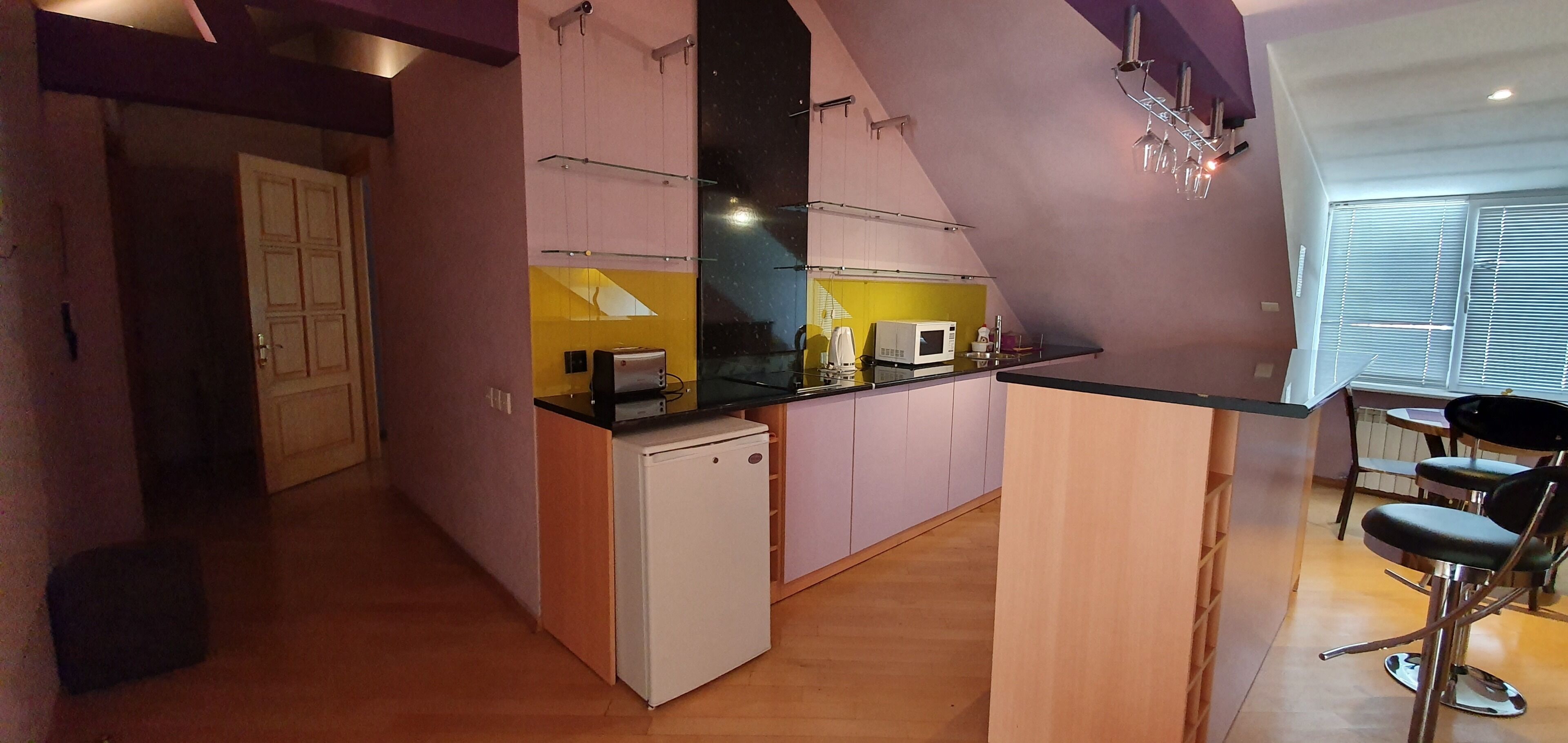 A compact kitchen with a microwave, toaster, and small refrigerator. There is a bar area with stools and a window with blinds.
