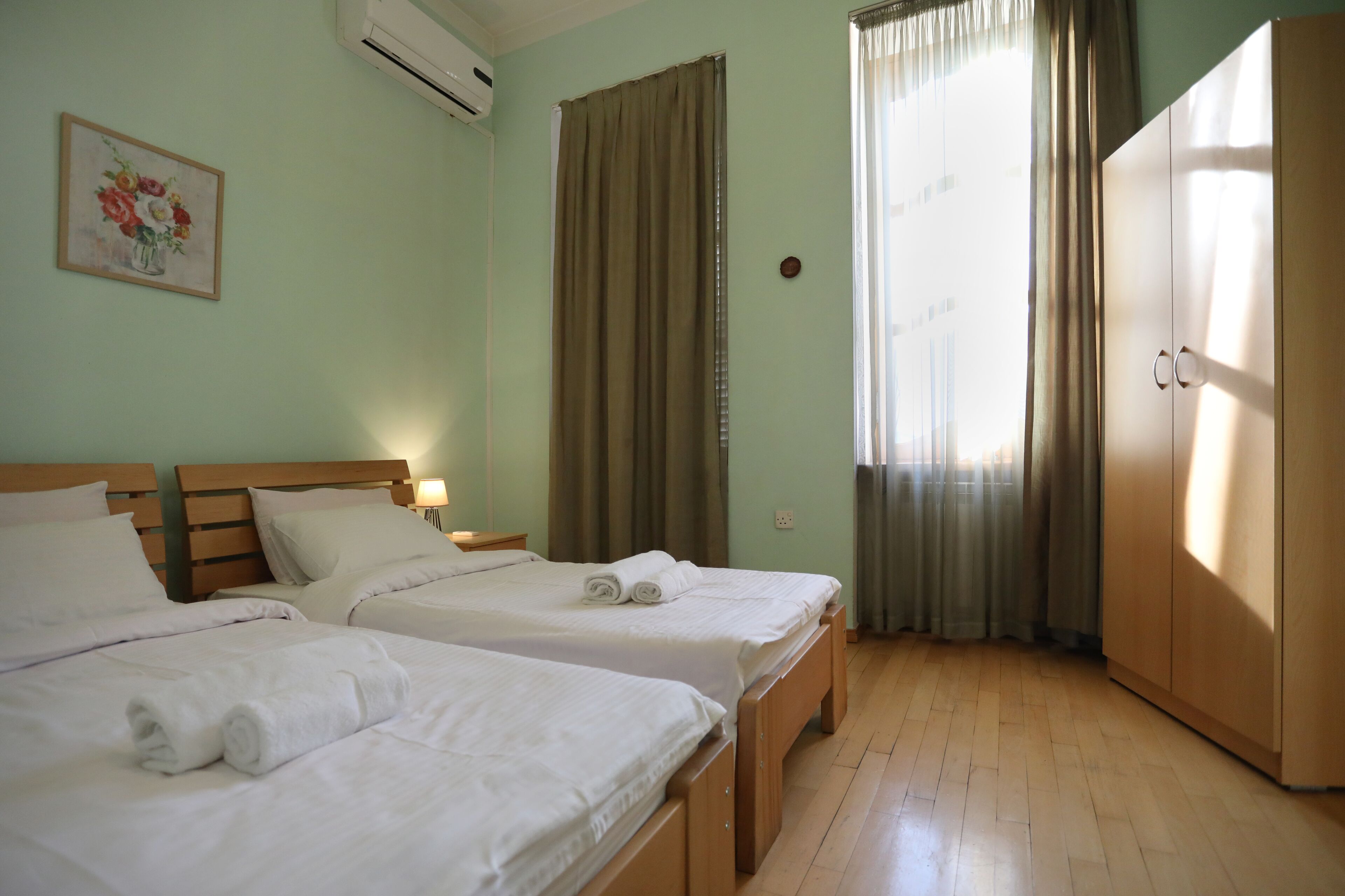 A hotel room with two beds, a wardrobe, a painting on the wall, and a window with curtains.