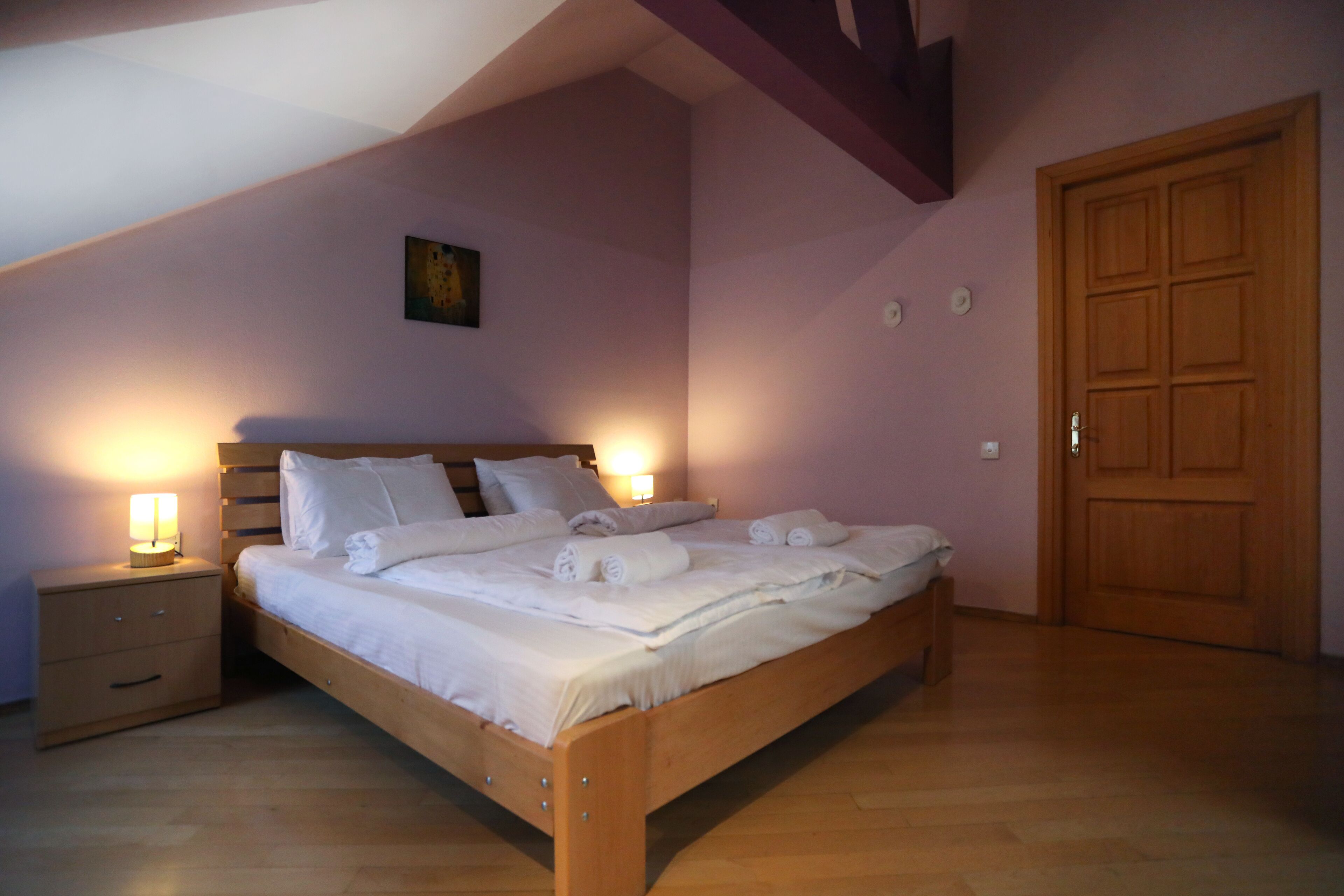A neatly made bed with white linens and rolled towels, a wooden nightstand with a lamp, and a wooden door in a room with wooden flooring and a sloped ceiling.