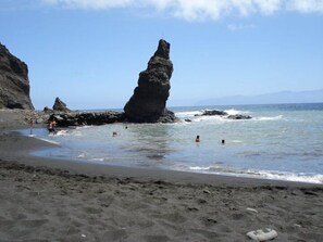 Una playa de arena oscura, una imponente formación rocosa y mar en calma.
