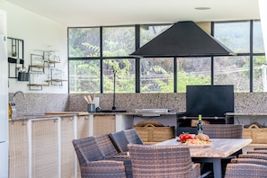Une cuisine moderne avec de grandes fenêtres, un barbecue, des meubles en osier et une table à manger où sont servis des plats.