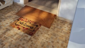 Un pasillo con una puerta de madera, un felpudo, una zona de comedor con mesa y sillas, y un mueble pequeño.