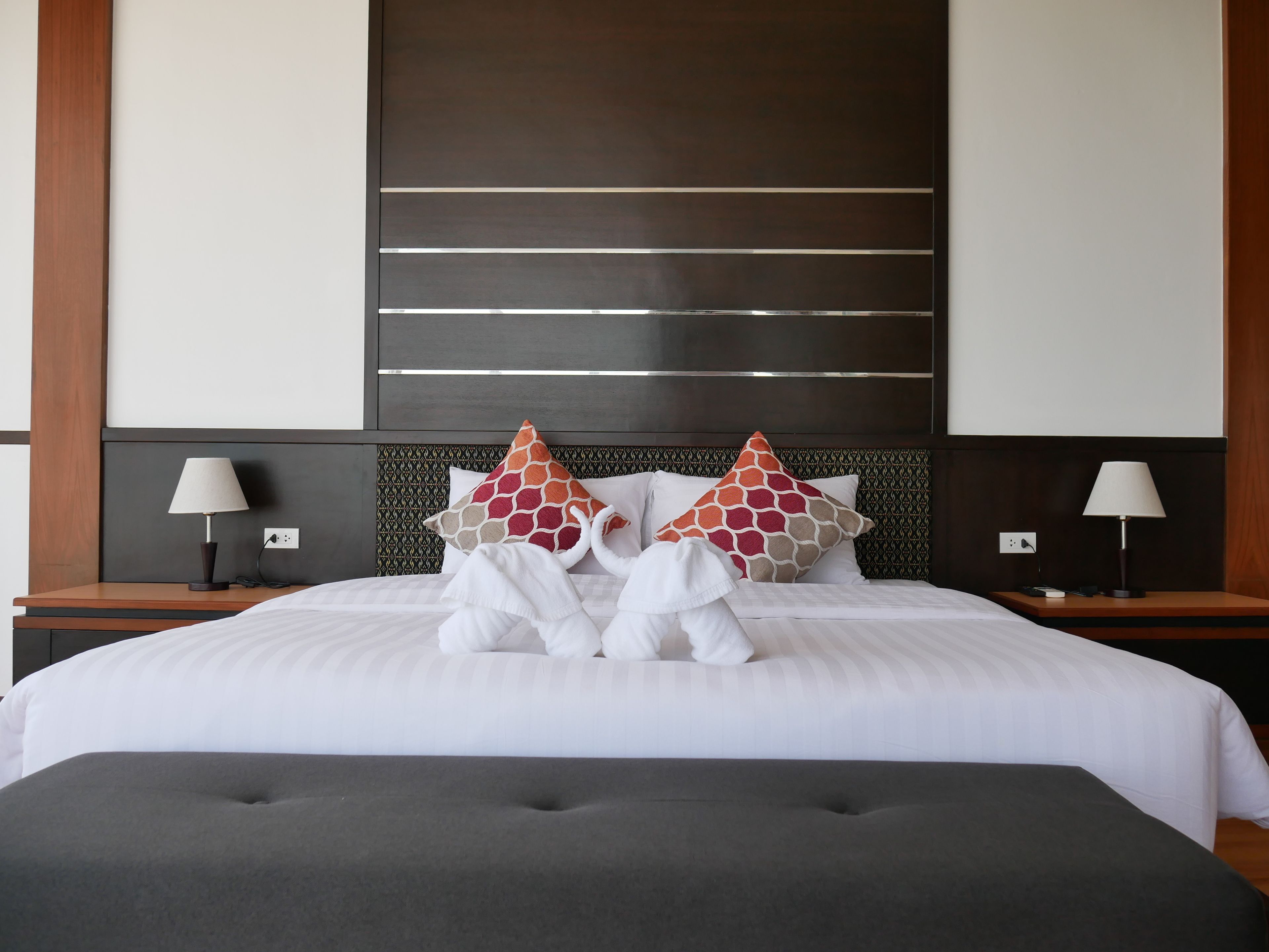 A neatly made bed with white linens, two decorative pillows, and a grey ottoman.