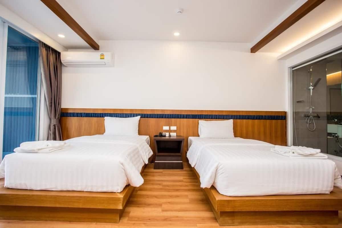 Habitación de hotel con dos camas, cabecero de madera, mesita de noche, unidad de aire acondicionado y zona de ducha con mampara de vidrio.