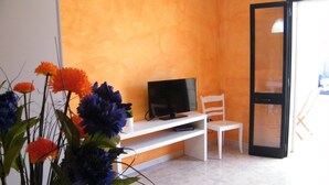 A living room with a television on a white stand, a white chair, and a colorful rug. - PINK BAY HOUSE 100 MT FROM THE SEA  (Marina San Gregorio)