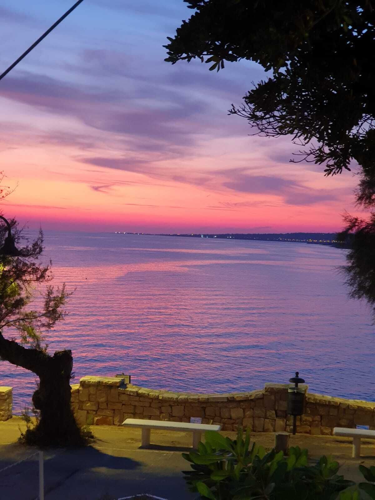 Una vista del atardecer sobre un mar tranquilo, con un muro de piedra y bancos a lo largo de la orilla.
