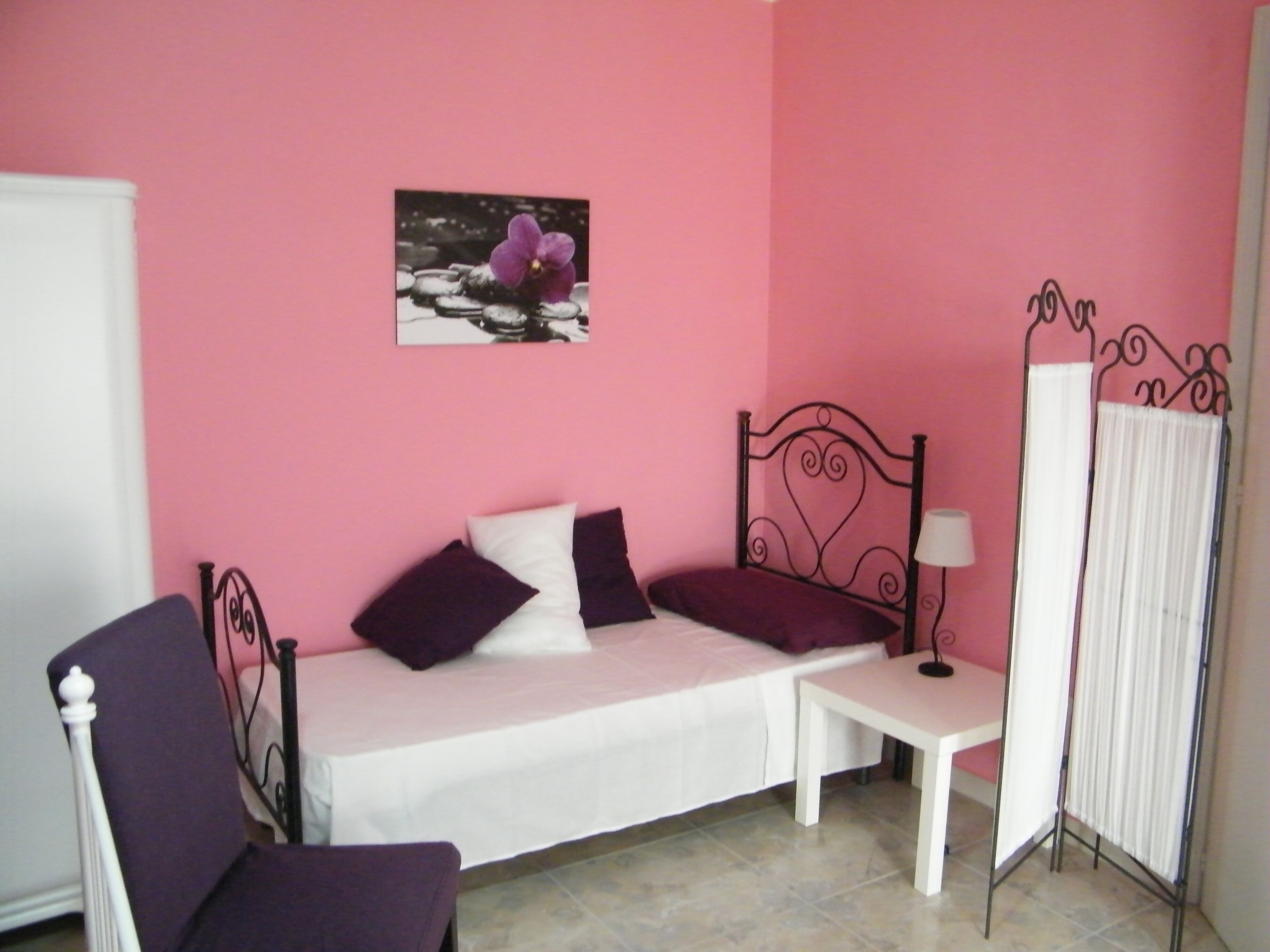 Une pièce avec un mur rose, un lit avec une literie blanche et violette, une petite table avec une lampe et une chaise.