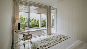 Un dormitorio con una cama grande, un escritorio y una silla. Se ve una ventana con cortinas y vistas al exterior con vegetación.