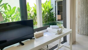 Una habitación moderna con una televisión de pantalla plana sobre una mesa blanca, una planta pequeña y toallas dobladas con cuidado.