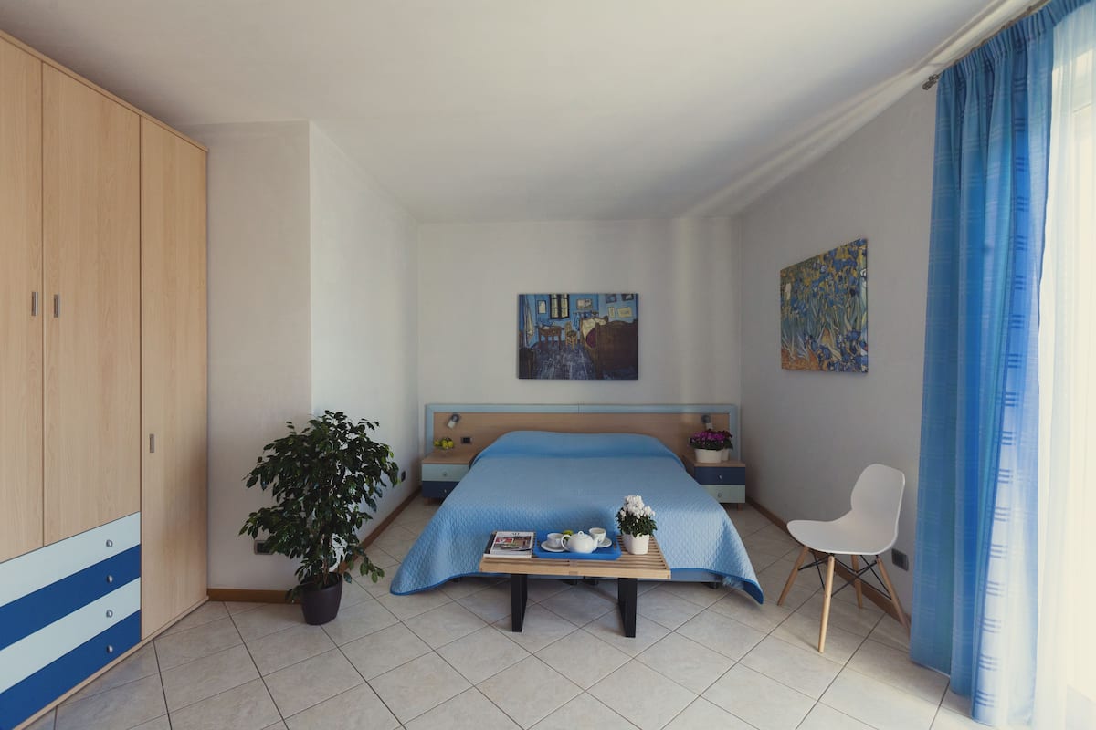 A bedroom with a bed, a bedside table, a chair, a potted plant, and a painting on the wall.