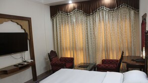 Habitación de hotel con una cama grande, dos sillones, una mesita, un televisor montado en un estante de madera y una cortina estampada.