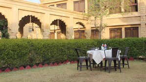 Un espacio de comedor al aire libre con una mesa y sillas, rodeado por un seto y un edificio con arcos.
