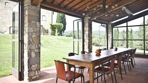 Un espace extérieur abrité, aménagé pour les repas, avec des piliers en pierre, une longue table en bois et des chaises.