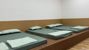 A row of four single beds with green bedding and pillows, arranged in a row against a wooden wall in a room with a wooden floor.