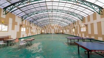 An indoor sports hall with a green floor, multiple ping pong tables, and a high ceiling with a metal framework.
