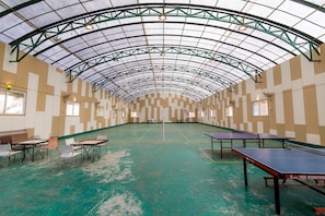An indoor sports hall with a green floor, multiple ping pong tables, and a high ceiling with a metal framework.