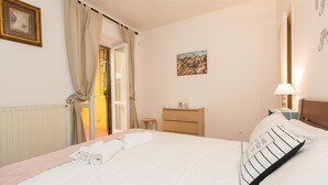 A hotel room with a bed, a nightstand, a small table, a chair, a radiator, a window with curtains, and a painting on the wall.