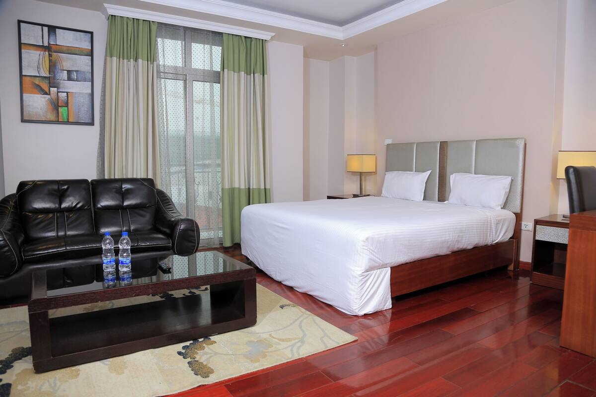 A hotel room with a bed, a leather sofa, a coffee table, and a nightstand with a lamp.