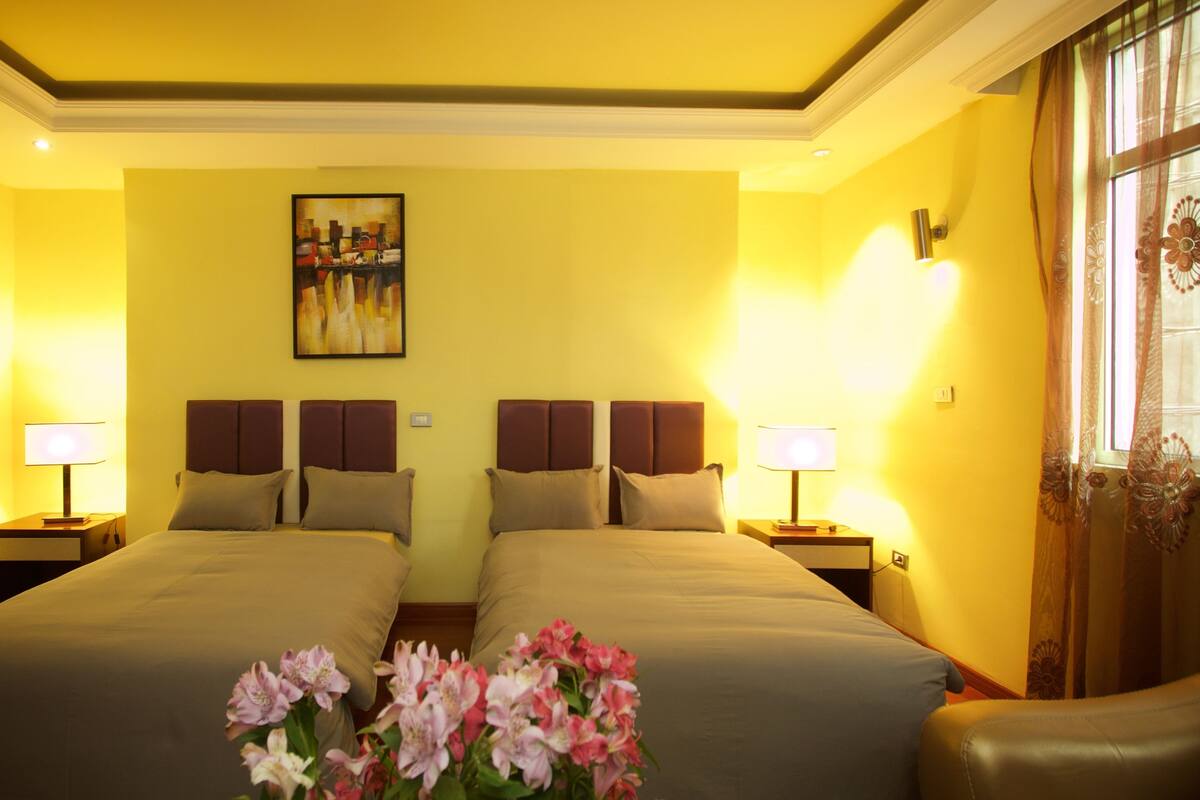 A hotel room with two beds, a painting on the wall, and a window with curtains.