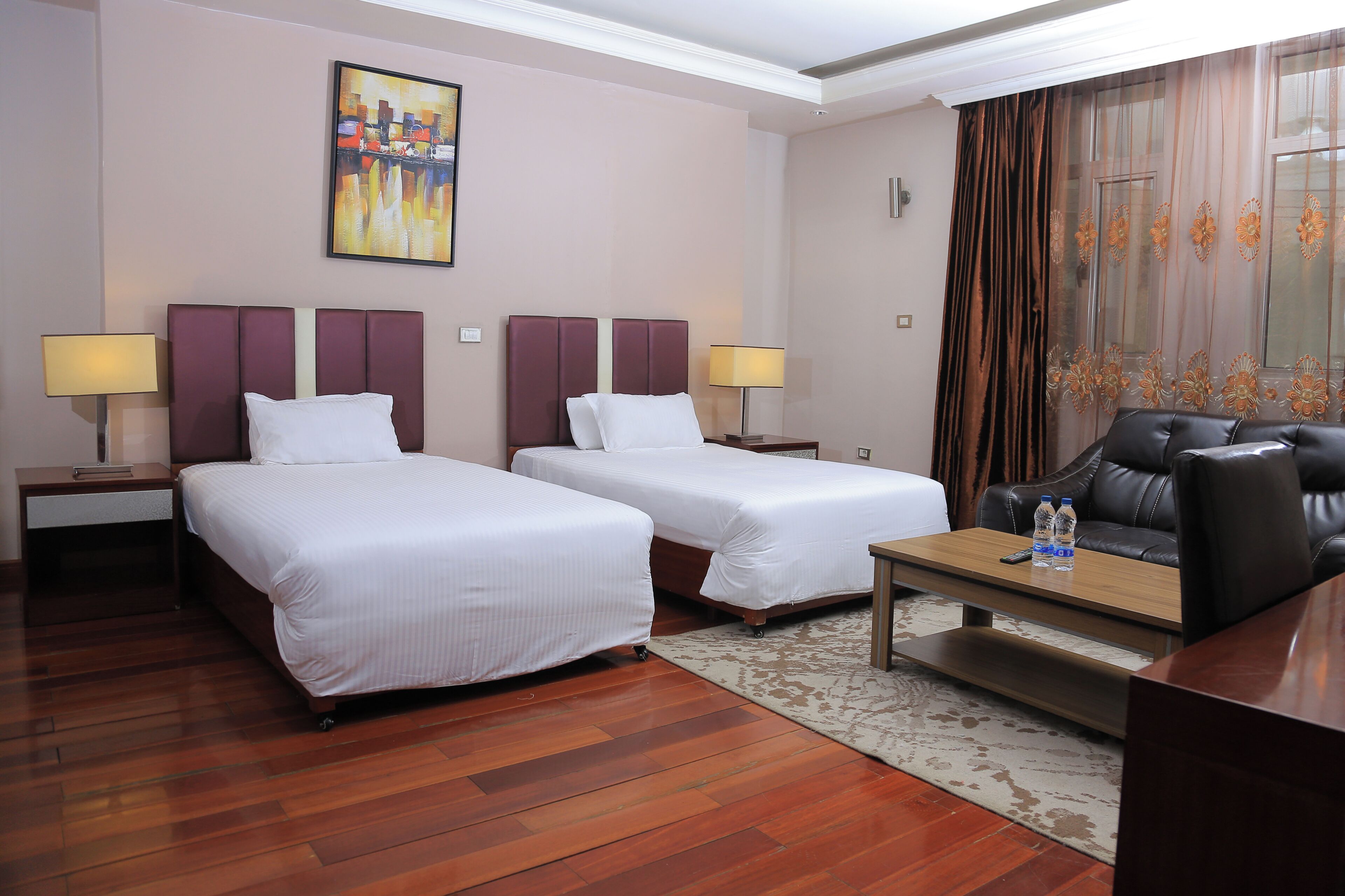 A hotel room with two beds, a desk, a chair, a lamp, and a painting on the wall.