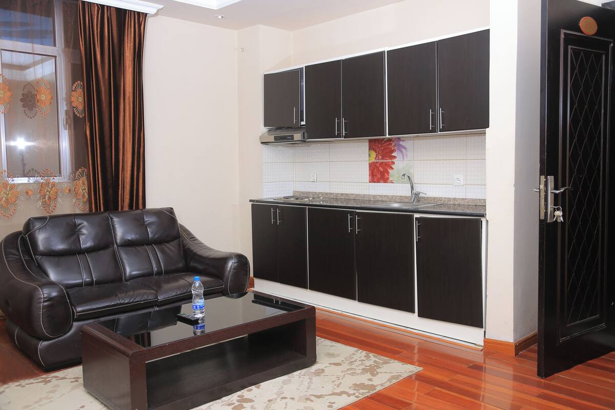 A living room with a leather sofa, a glass coffee table, and a kitchen area with black cabinets and a tiled backsplash.