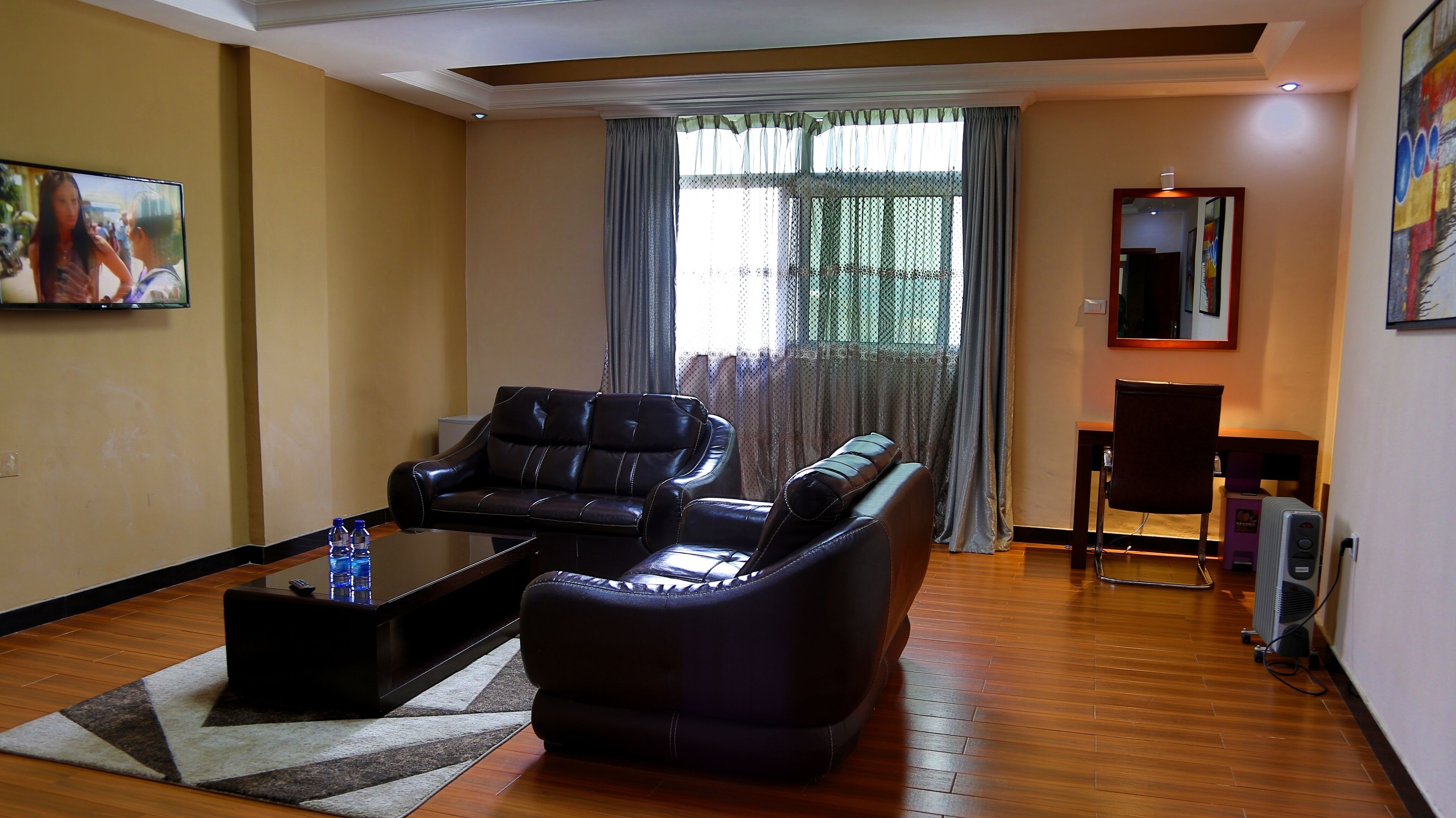 A living room with a leather sofa, a coffee table, and a TV. There is a window with curtains and a painting on the wall.
