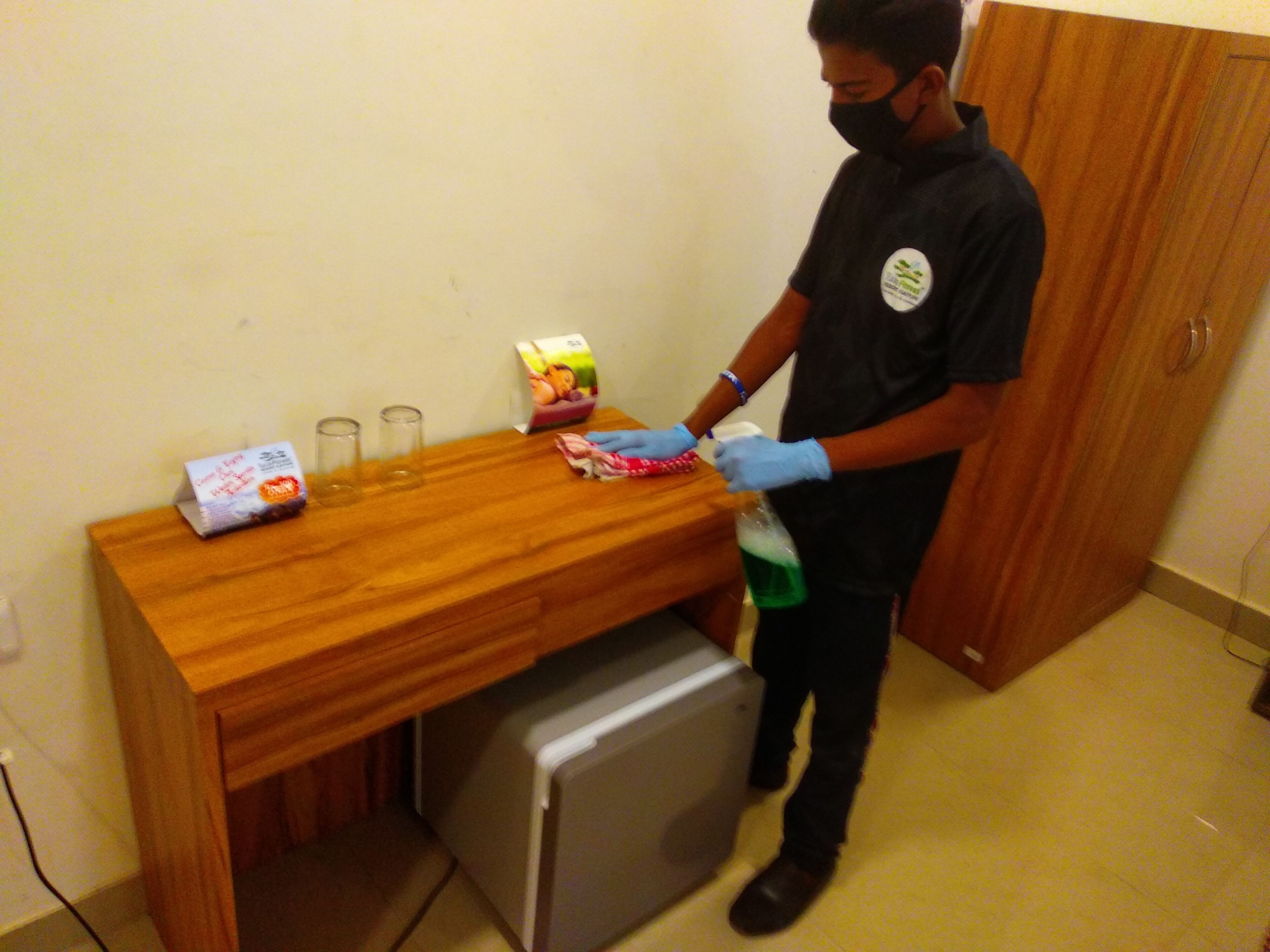 A person cleaning a wooden table with a spray bottle and cloth, a box of tissues, and two glasses on the table.