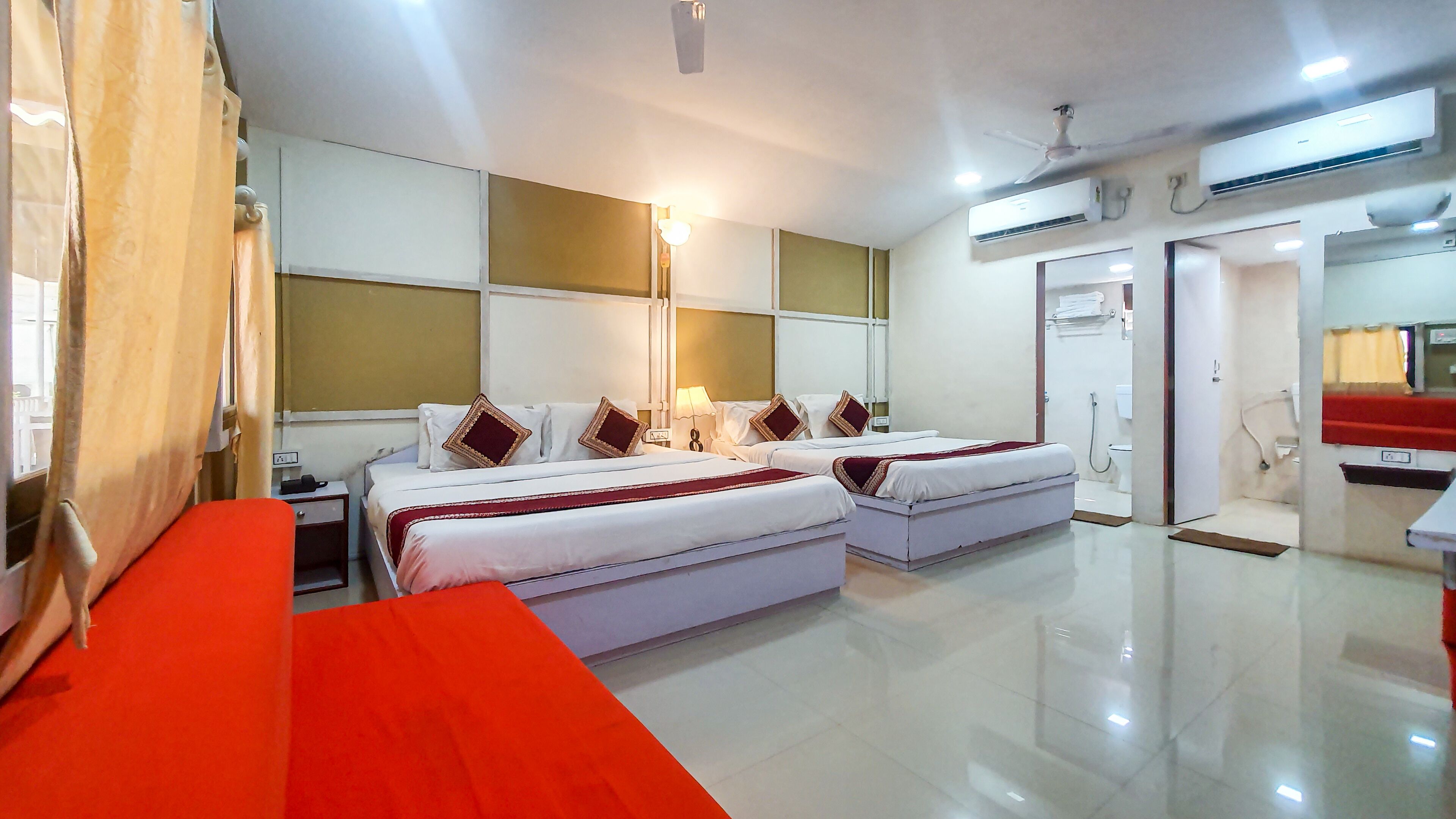 Une chambre d’hôtel avec deux lits, un banc rouge, une salle de bain avec douche et un appareil de climatisation.