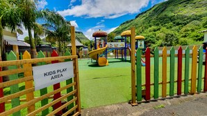 Zona de juegos con tobogán y estructuras para escalar, rodeada por una cerca y follaje.