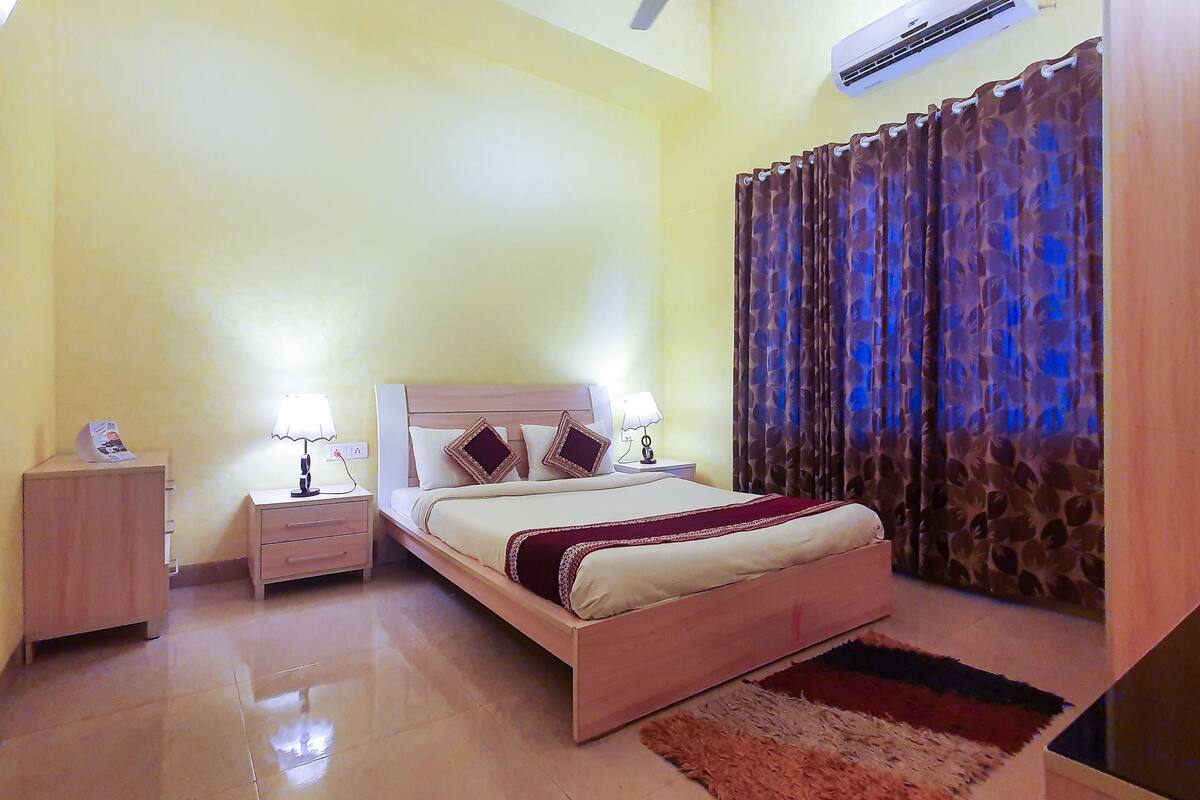 A hotel room with a bed, bedside tables, a nightstand, a lamp, a curtain, and a wall-mounted air conditioner.