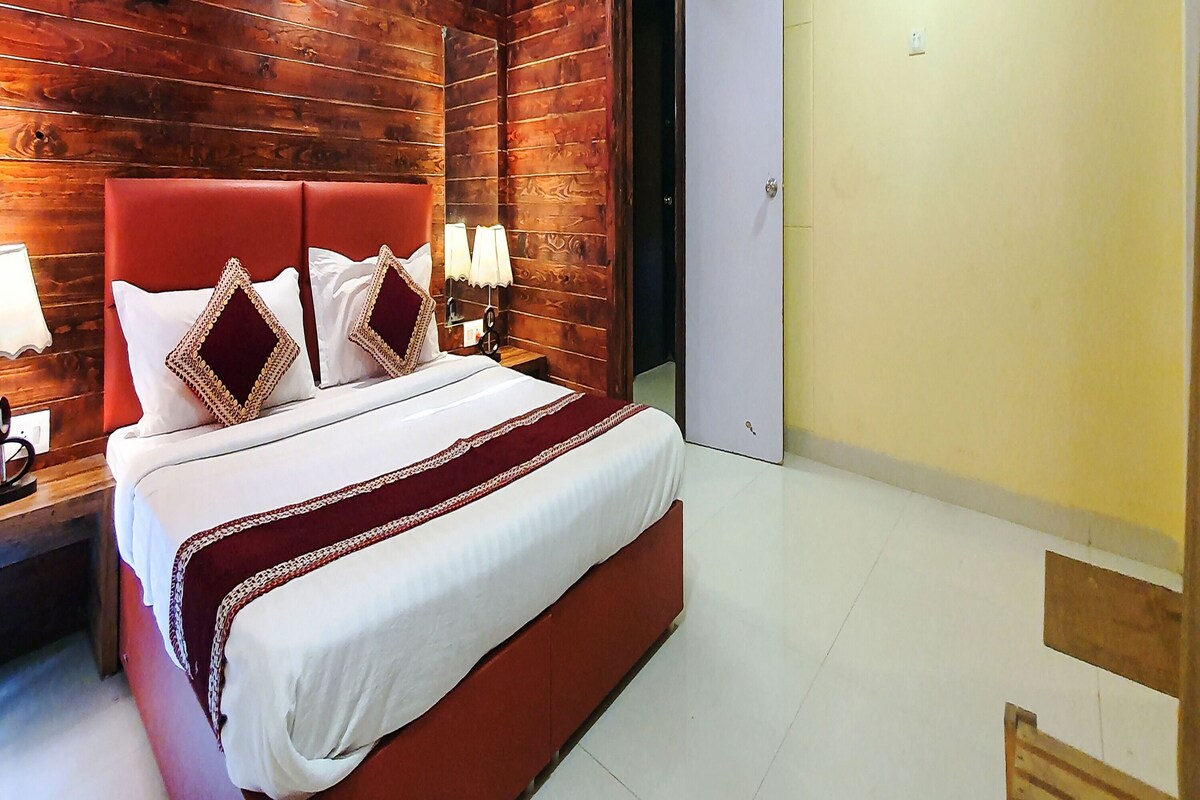 Quarto de hotel com parede de madeira, cama com roupa de cama branca e detalhes vermelhos, duas luminárias de cabeceira e uma porta.