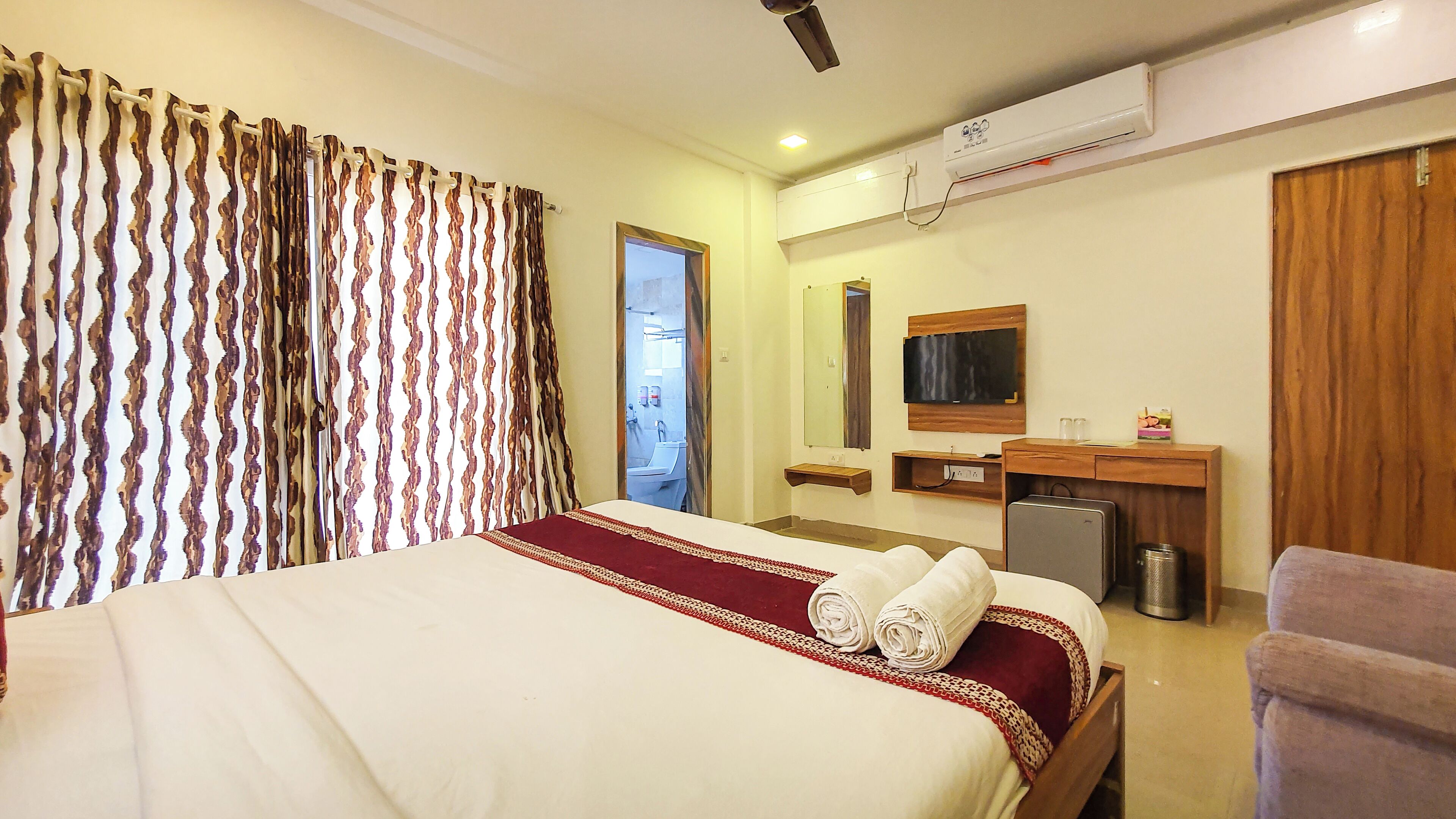 A hotel room with a bed, a desk, a mirror, a TV, and an air conditioning unit.