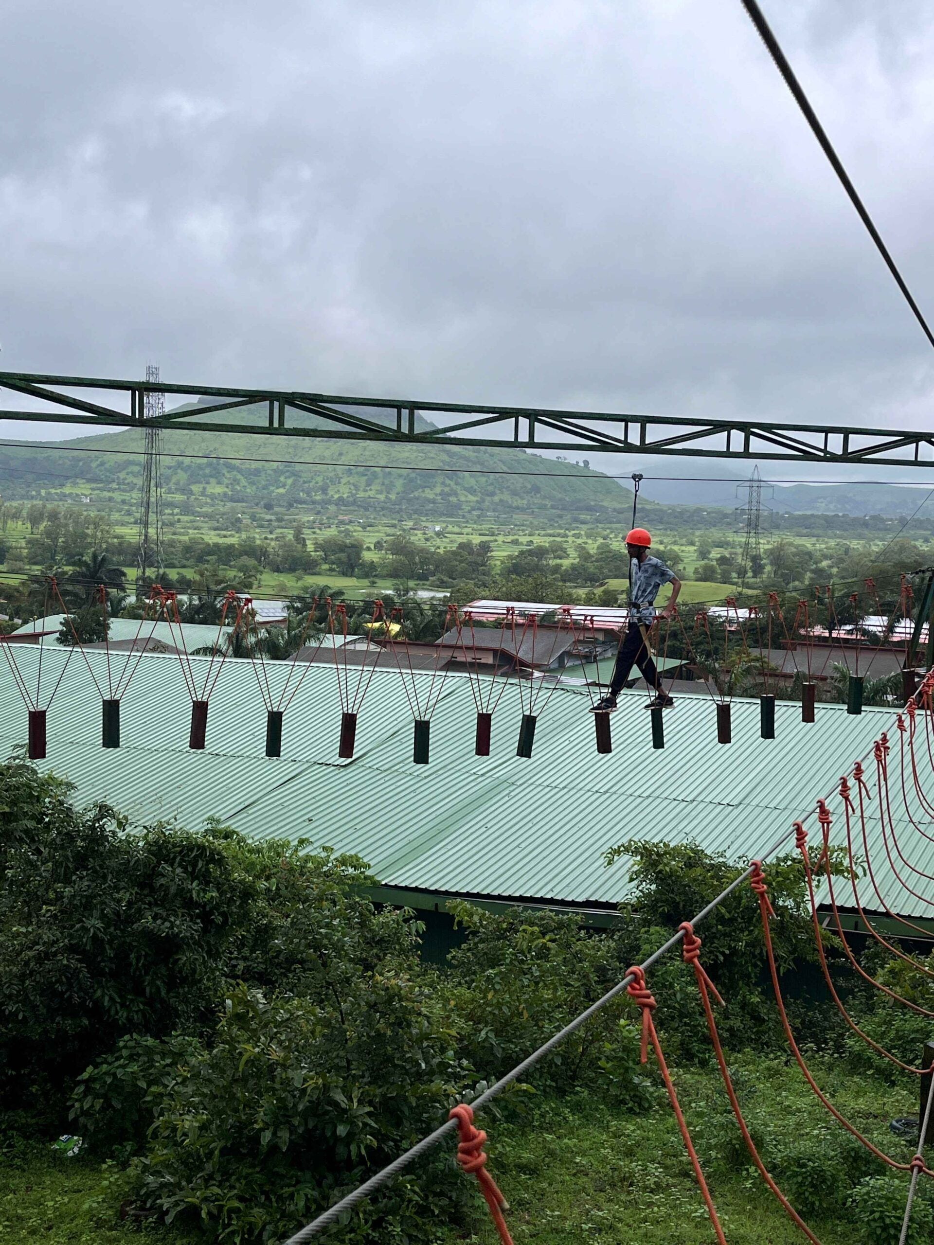 A person walking on a suspended bridge with a green roof and red ropes, overlooking a green landscape with hills and power lines.