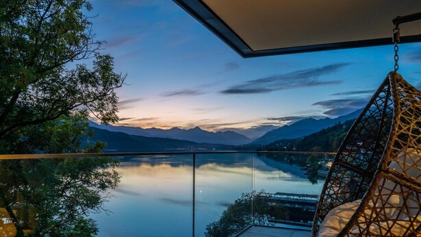 Un balcón con una hamaca colgante con vistas a un lago tranquilo y montañas al anochecer.