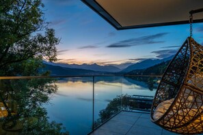 A balcony with a hanging chair overlooking a calm lake and mountains at dusk. - Villa Postillion am See (Millstatt)