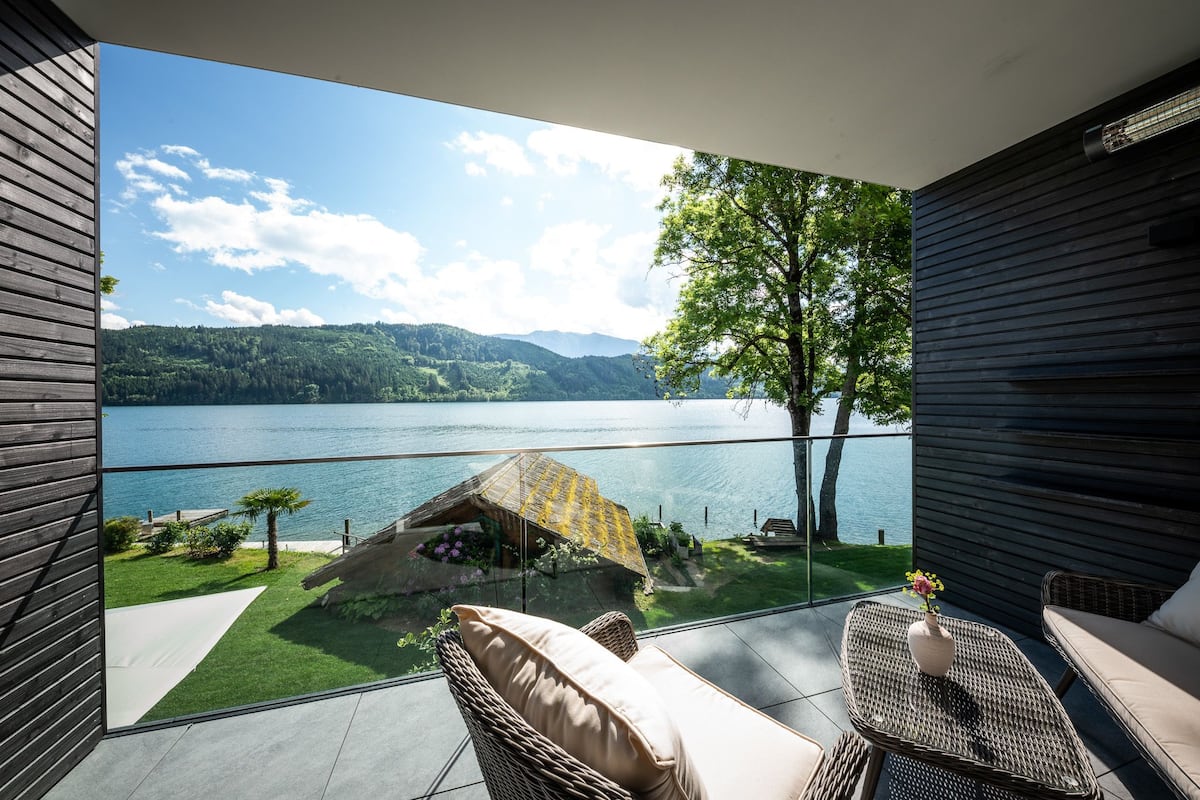 Un balcon doté de meubles en osier, d’une porte vitrée et offrant une vue sur un lac et des montagnes.