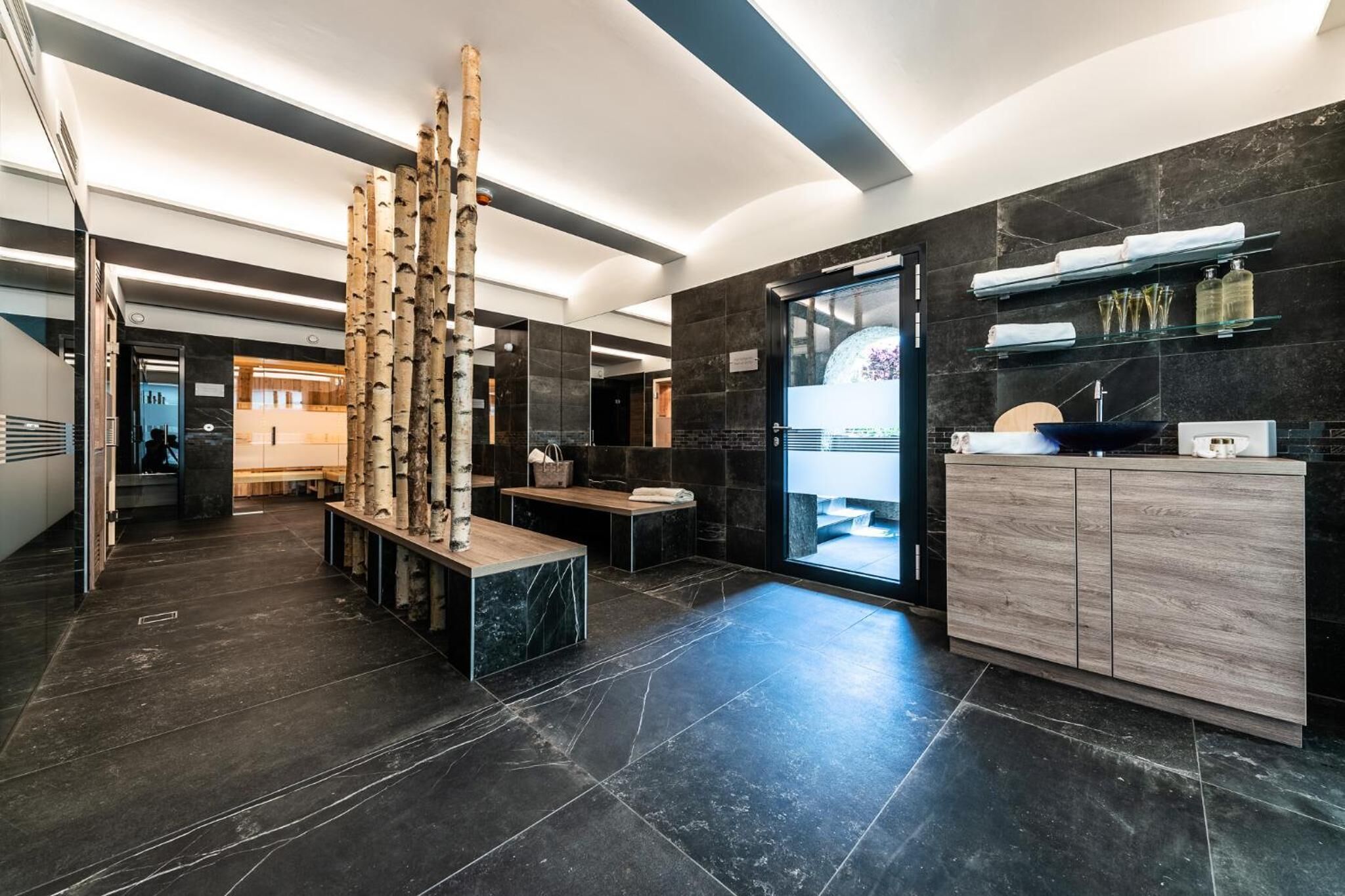 Un baño moderno con baldosas de mármol oscuro, un mueble de tocador de madera y un box de ducha de cristal.