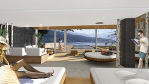 Habitación de hotel moderna con un gran ventanal que da a un lago y montañas. Hay dos sillones, una mesita y un área circular con cojines.