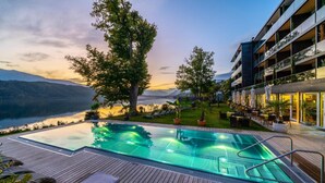 Un hotel moderno con piscina infinita en la azotea, con vistas a un lago, rodeado de exuberante vegetación y montañas al fondo.