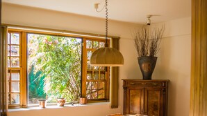 Un coin repas avec une table et des chaises en bois, un luminaire suspendu, une fenêtre donnant sur un espace verdoyant et un meuble agrémenté d’une plante.