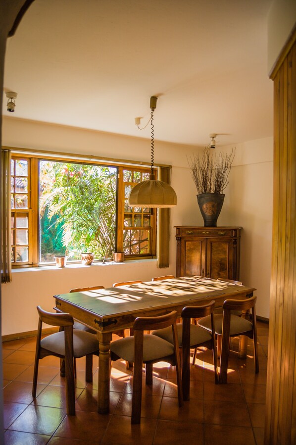 A dining area with a wooden table and chairs, a hanging light fixture, a window with a view of greenery, and a cabinet with a plant.