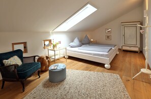 Ein gemütliches Schlafzimmer mit schrägem Dach, einem Bett mit Kissen, einem Sessel, einem kleinen Tisch mit einer Vase und einem Schrank.