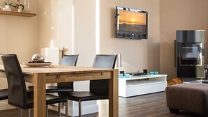 Un salon moderne avec une table à manger en bois, des chaises en cuir noir, un téléviseur à écran plat fixé au mur, un coin cuisine intégré avec un réfrigérateur en acier inoxydable et un coin salon confortable avec un pouf marron.