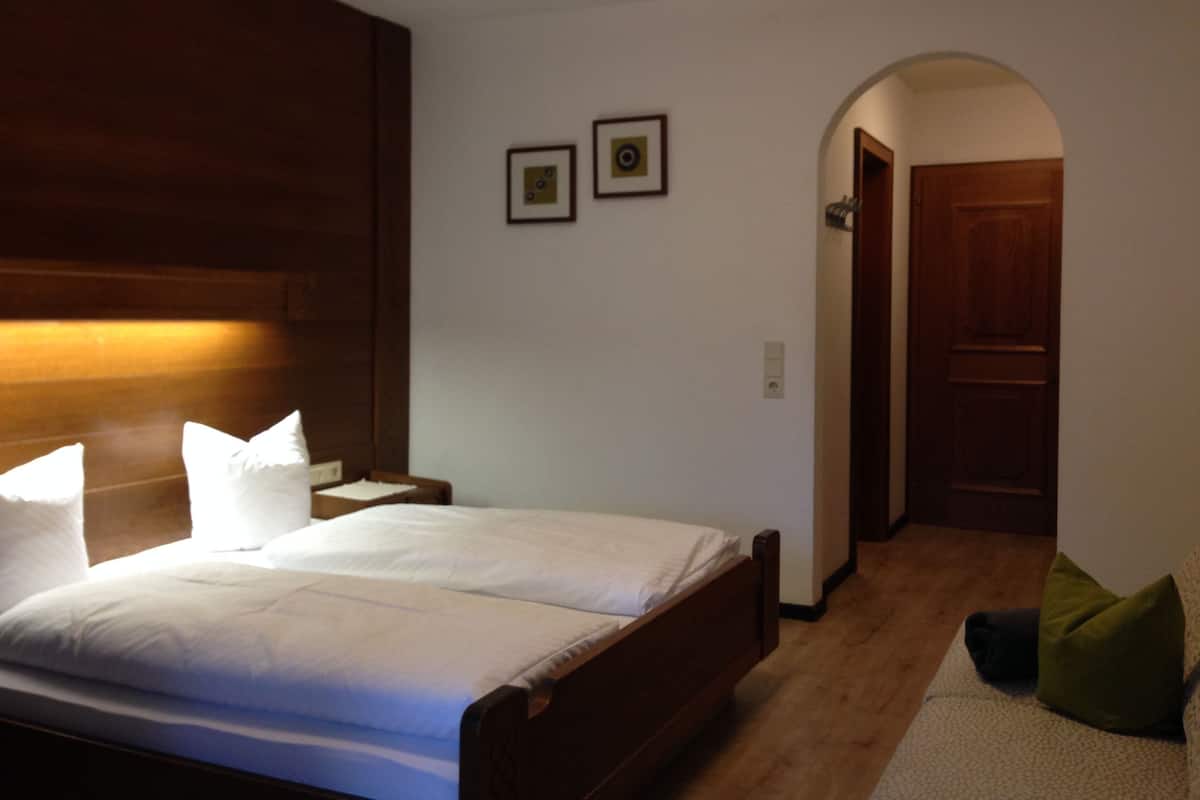 A bedroom with a wooden headboard, a bed with white linens, two framed pictures on the wall, and a wooden door in the background.